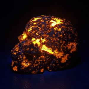 May include: A glowing skull sculpture with a dark background. The skull is made of a dark material with orange and yellow highlights, creating a striking contrast. The sculpture is a unique decorative item.