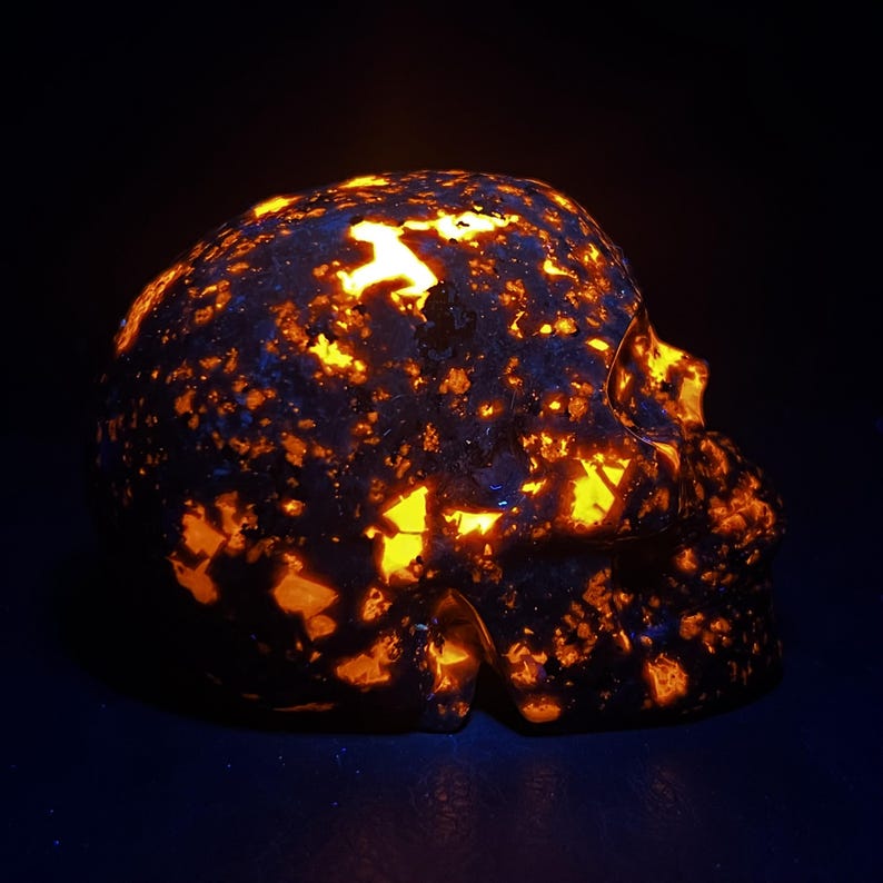 May include: A skull-shaped sculpture with a dark, textured surface, illuminated by vibrant orange and yellow light. The contrast between the dark base and the glowing highlights creates a striking visual effect. The sculpture is a unique decorative item.