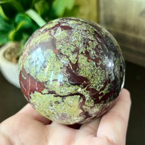 Puede incluir: Una esfera pulida de jaspe de sangre de dragón, que muestra una mezcla de tonos rojo intenso y verde. La superficie de la piedra es lisa y reflectante, con un aspecto veteado. Esta esfera decorativa es una adición única a cualquier colección.