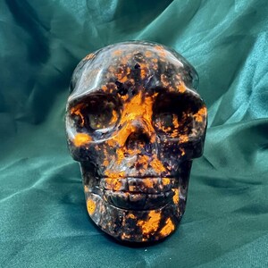 May include: A decorative skull sculpture with a dark base color and vibrant orange accents. The skull's features are clearly defined, and the overall design has a unique, artistic appearance. The sculpture is likely made of a polished stone or resin material.