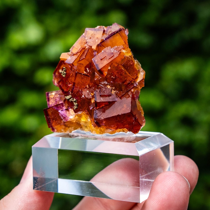 May include: A cluster of translucent, deep orange and amber-coloured fluorite crystals with visible cubic formations and small metallic inclusions, mounted on a clear, rectangular acrylic display base for stability and viewing.