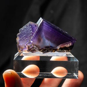 Puede incluir: Un vibrante espécimen de cristal de fluorita púrpura con forma geométrica. El cristal se encuentra sobre un expositor acrílico transparente. La fluorita tiene una apariencia en capas con una mezcla de tonos púrpuras oscuros y claros. El fondo es negro.