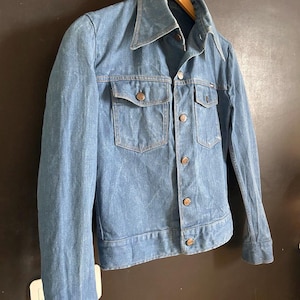May include: A light blue denim jacket with a wide collar and two chest pockets. The jacket has a button-up front and buttoned cuffs. The jacket is hanging on a wooden hanger against a dark background. The jacket appears to be vintage.
