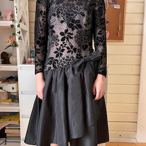 May include: A black dress with a sheer, floral-patterned bodice and long sleeves. The dress features a satin skirt with a bow detail at the waist. The dress has a vintage style.