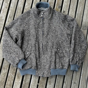 May include: A black and white speckled tweed bomber jacket featuring a high collar with two buttons, a front zipper closure, two side pockets with button flaps, and solid grey ribbed knit cuffs and a matching ribbed hem at the waist.