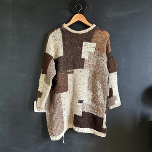 May include: A long-sleeved sweater with a patchwork design in shades of brown, beige, and tan hangs on a wooden hanger. The sweater has a crew neck and a loose, oversized fit. The fabric appears to be a chunky knit.