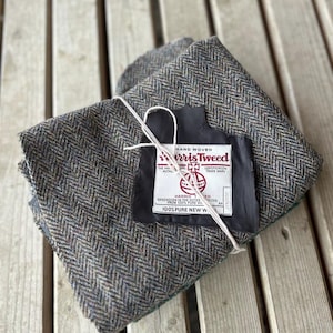 May include: A folded piece of grey and brown herringbone patterned Harris Tweed fabric. A white rectangular label is attached with white string, featuring the text Harris Tweed, Hand Woven, Certification Trade Mark, and 100% Pure New Wool.