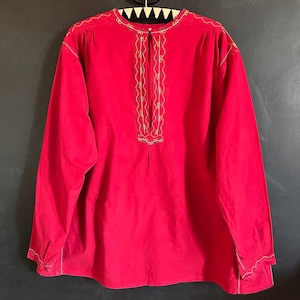 May include: A long-sleeved, red velvet shirt with gold-colored embroidered detailing. The embroidery is featured along the neckline and down the front of the shirt. The shirt is hanging on a black hanger against a dark background.