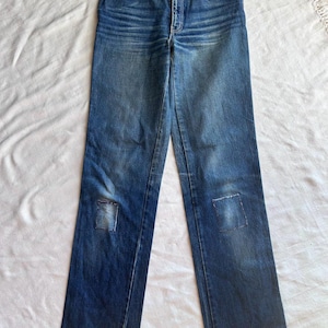 May include: Blue denim jeans with a high waist and straight leg. The jeans feature a classic five-pocket design and a button and zip closure. Patches are sewn on the knees. The jeans are laid flat on a white surface.