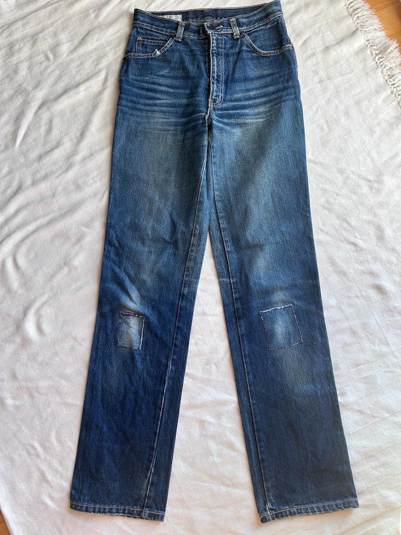 May include: Blue denim jeans with a high waist and straight leg. The jeans feature a classic five-pocket design and a button and zip closure. Patches are sewn on the knees. The jeans are laid flat on a white surface.