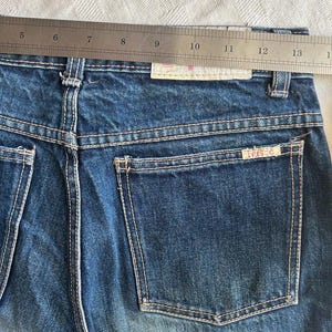 May include: Close-up of blue denim jeans with a back pocket and white stitching. A metal ruler is placed across the waistband. The jeans have a faded appearance and a small label with the text "trisse" is visible on the back pocket.
