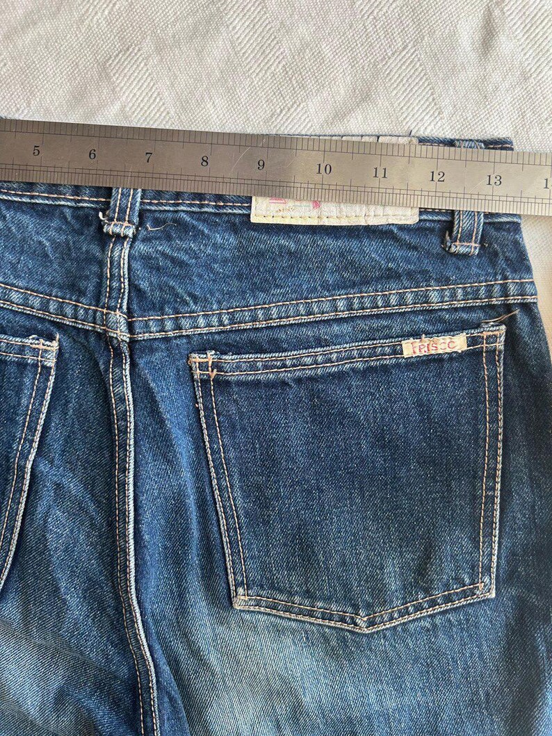 May include: Close-up of blue denim jeans with a back pocket and white stitching. A metal ruler is placed across the waistband. The jeans have a faded appearance and a small label with the text "trisse" is visible on the back pocket.