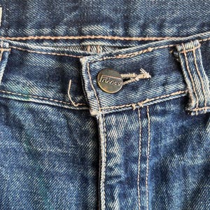 May include: Close-up of blue denim jeans. The jeans have a button closure with the brand name "Trinco" and visible stitching. The denim has a faded, worn appearance.