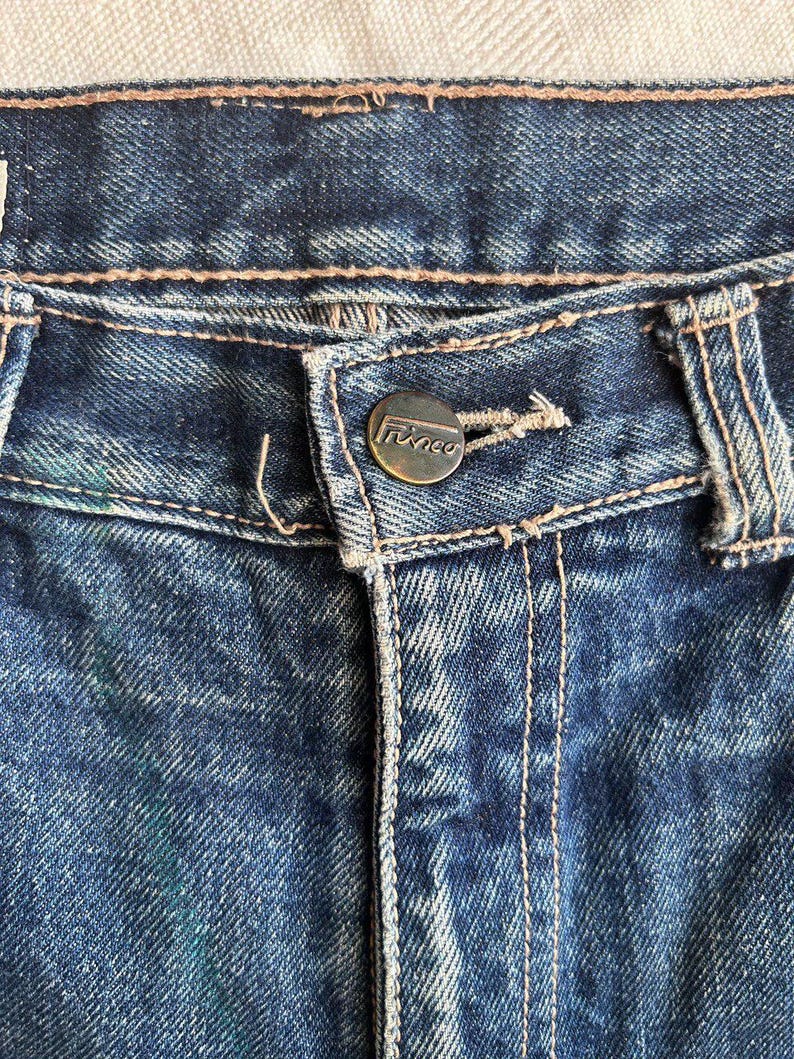 May include: Close-up of blue denim jeans. The jeans have a button closure with the brand name "Trinco" and visible stitching. The denim has a faded, worn appearance.