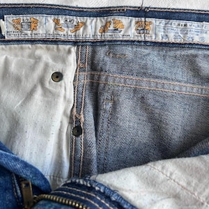 May include: Close-up of blue denim jeans with a button closure and zipper. The jeans have a white interior and a label with cartoon bear illustrations and text. The label reads "HENNES H&M MAURITZ 100% BOMULL 100% BAUMWOLLE 100% COTTON 100% COTON."