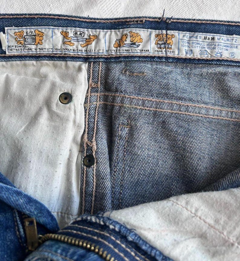 May include: Close-up of blue denim jeans with a button closure and zipper. The jeans have a white interior and a label with cartoon bear illustrations and text. The label reads "HENNES H&M MAURITZ 100% BOMULL 100% BAUMWOLLE 100% COTTON 100% COTON."