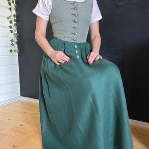 Op de afbeelding: Een groen en wit geruit lijfje jurk met zilveren knopen en een lange groene rok. De jurk wordt gecombineerd met een wit shirt met korte mouwen. De jurk is een traditionele stijl.