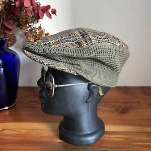May include: A flat cap featuring a multi-colored plaid pattern on the crown and a solid olive green knit band around the base. The cap is displayed on a black mannequin head wearing gold-rimmed aviator sunglasses with dark lenses.