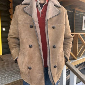 May include: A tan shearling coat with a double-breasted design. The coat features dark brown buttons, a wide collar, and a light brown interior lining. The coat has two front pockets and dark brown trim.