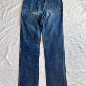 May include: Blue denim jeans with a straight leg cut. The jeans have two back pockets and a small label at the back. The denim has a faded appearance, with lighter areas on the thighs and seat. The jeans are laid flat on a white surface.