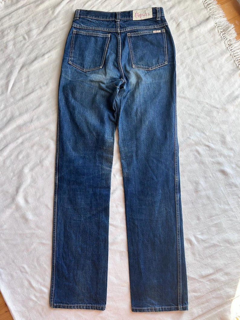 May include: Blue denim jeans with a straight leg cut. The jeans have two back pockets and a small label at the back. The denim has a faded appearance, with lighter areas on the thighs and seat. The jeans are laid flat on a white surface.