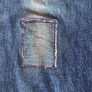 May include: Close-up of blue denim fabric with a rectangular patch sewn on. The patch is a lighter shade of blue and is outlined with a dark purple stitch. The denim has a worn appearance.
