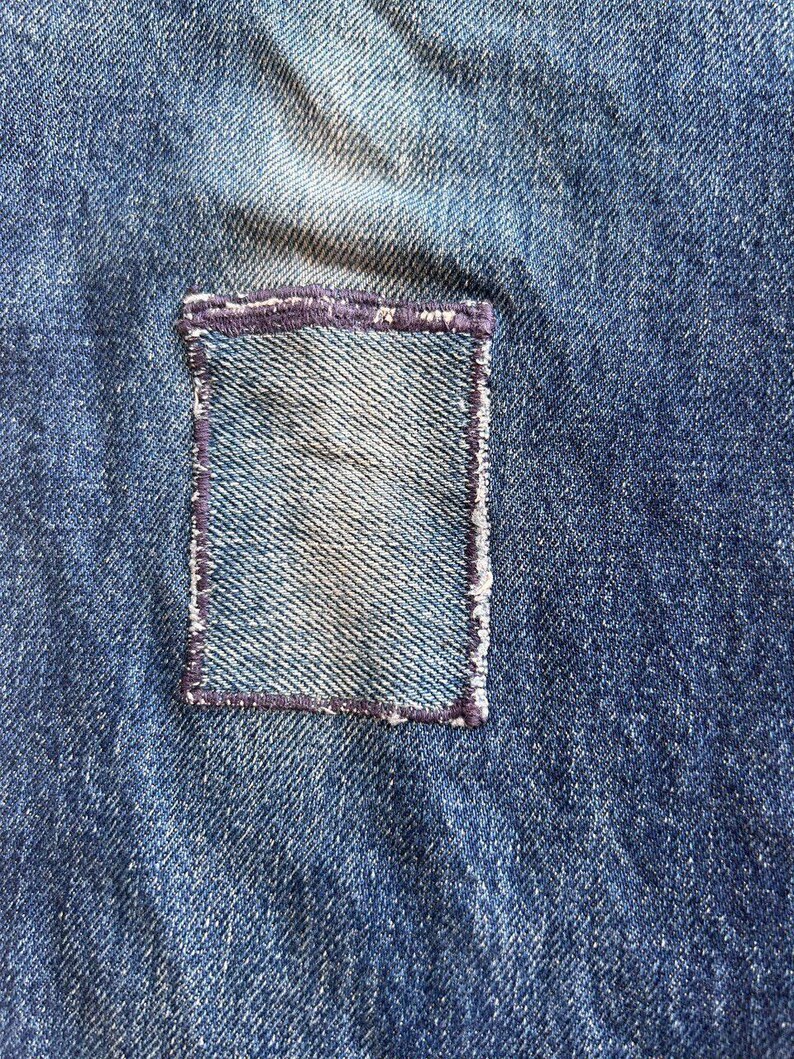 May include: Close-up of blue denim fabric with a rectangular patch sewn on. The patch is a lighter shade of blue and is outlined with a dark purple stitch. The denim has a worn appearance.