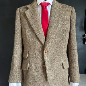 May include: A tan and brown herringbone blazer with two front buttons and two flap pockets, worn over a crisp white collared shirt and a bright red necktie. The jacket features notched lapels and long sleeves with white cuffs visible at the wrists.