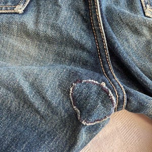 May include: Close-up of blue denim jeans with a visible patch repair. The patch is a lighter shade of blue and is stitched with a contrasting thread. The jeans show signs of wear and tear.