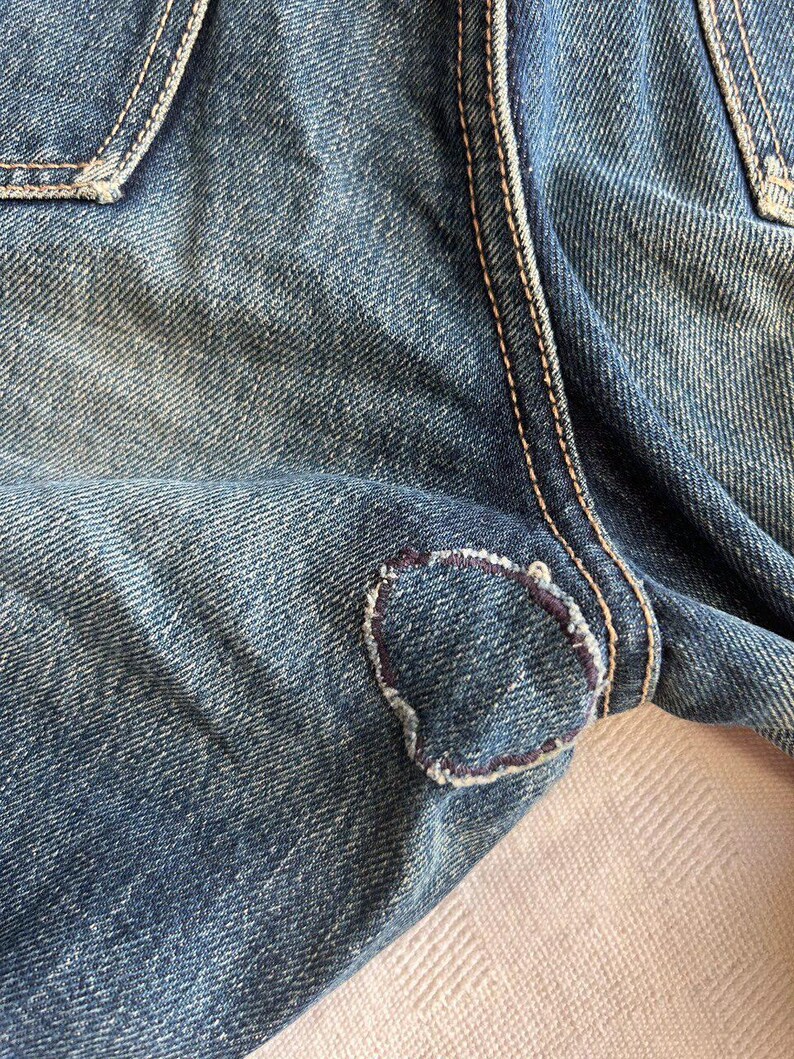 May include: Close-up of blue denim jeans with a visible patch repair. The patch is a lighter shade of blue and is stitched with a contrasting thread. The jeans show signs of wear and tear.