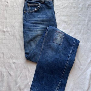 May include: Blue denim jeans with a faded wash and a patch detail on the back pocket. The jeans are folded and laid flat on a white surface, showcasing the classic five-pocket design and a button and zip closure.
