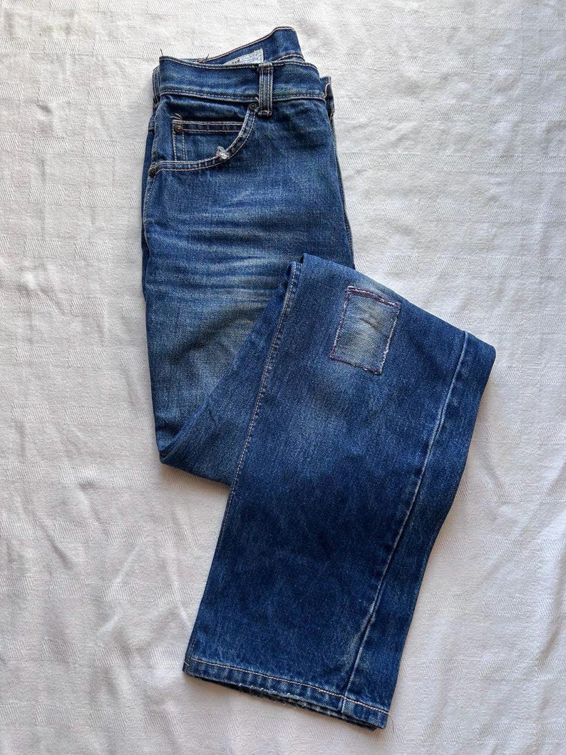 May include: Blue denim jeans with a faded wash and a patch detail on the back pocket. The jeans are folded and laid flat on a white surface, showcasing the classic five-pocket design and a button and zip closure.