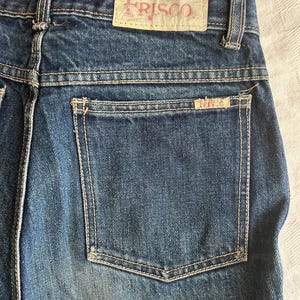 May include: Close-up of blue denim jeans with a back pocket and a "FRISCO" label. The jeans have light-coloured stitching and a faded appearance. The label is a light tan colour with the word "FRISCO" in a pinkish-red font.