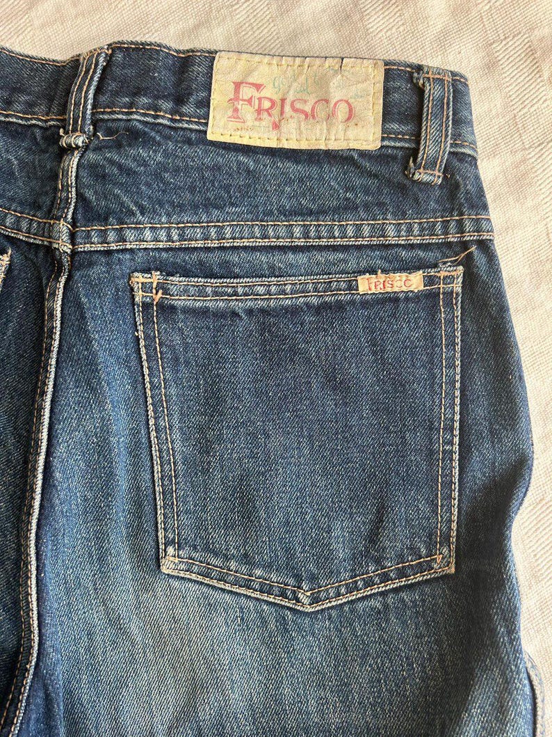 May include: Close-up of blue denim jeans with a back pocket and a "FRISCO" label. The jeans have light-coloured stitching and a faded appearance. The label is a light tan colour with the word "FRISCO" in a pinkish-red font.