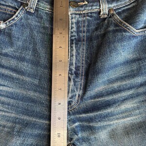 May include: Close-up of blue denim jeans with a metal ruler measuring the inseam. The ruler shows a measurement of approximately 30 cm. The jeans have a button closure and visible stitching details.