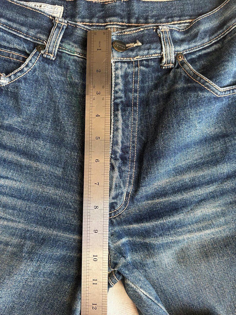 May include: Close-up of blue denim jeans with a metal ruler measuring the inseam. The ruler shows a measurement of approximately 30 cm. The jeans have a button closure and visible stitching details.