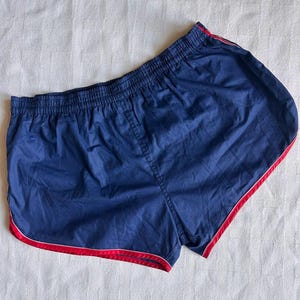 Puede incluir: Pantalones cortos deportivos azul marino con ribete rojo. Los pantalones cortos tienen cintura elástica y un diseño clásico. El ribete rojo perfila los bordes, añadiendo un toque de color a la tela azul oscuro.