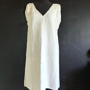 May include: An off-white linen dress with a V-neck, sleeveless design. The dress is displayed on a black mannequin against a dark background. The dress has a simple, classic design, suitable for casual wear.
