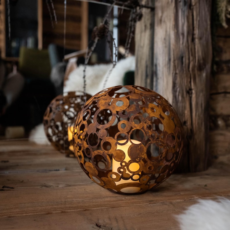 May include: Two spherical, rusty-brown metal lanterns with circular cutouts. The lanterns are lit from within, casting a warm glow. They sit on a wooden surface, with a rustic wooden beam in the background. The lanterns are decorative.