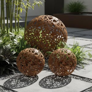May include: Three spherical, rusted metal garden ornaments. The spheres feature a geometric openwork design, revealing the greenery behind. They are arranged on a stone patio. The largest sphere is approximately 60 cm in diameter.