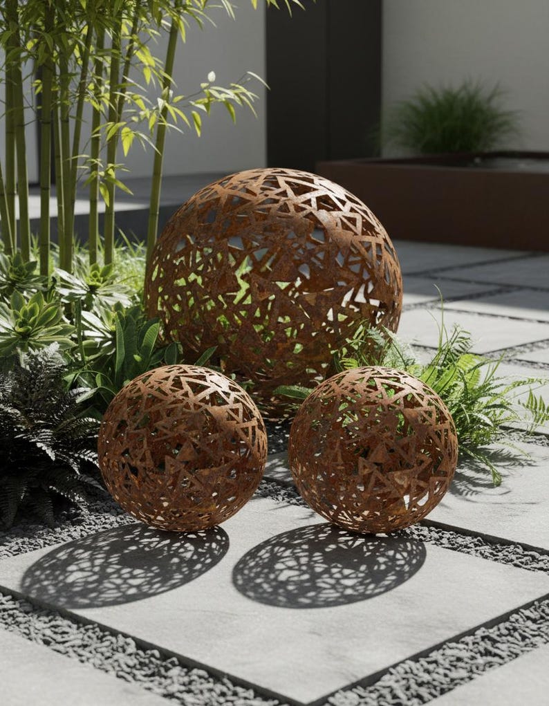 May include: Three spherical, rusted metal garden ornaments. The spheres feature a geometric openwork design, revealing the greenery behind. They are arranged on a stone patio. The largest sphere is approximately 60 cm in diameter.