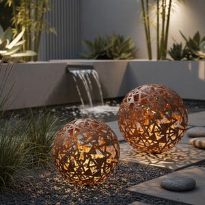 May include: Two spherical, rusty-orange metal garden lights with geometric cutouts. The lights are illuminated, casting warm light and shadows on the gravel and stone patio. The scene includes a small waterfall and lush greenery, creating a tranquil atmosphere.