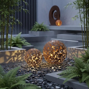 May include: Two spherical, rust-coloured metal garden lights with geometric cutouts, illuminated from within. They sit on a bed of dark pebbles, surrounded by ferns and bamboo, creating a tranquil garden scene.