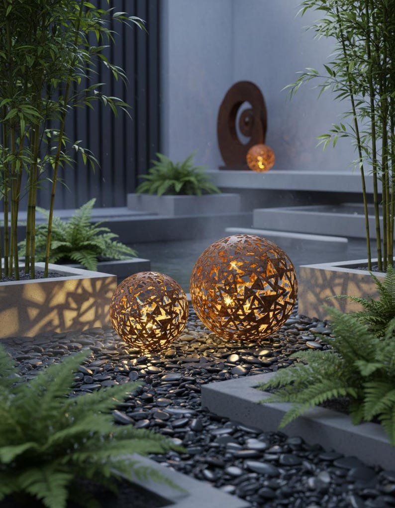 May include: Two spherical, rust-coloured metal garden lights with geometric cutouts, illuminated from within. They sit on a bed of dark pebbles, surrounded by ferns and bamboo, creating a tranquil garden scene.