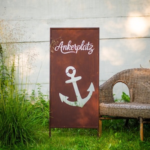 Könnte beinhalten: Ein verwittertes braunes Metallschild mit dem Wort "Ankerplatz" in weißer Schrift und einer weißen Ankergrafik. Das Schild befindet sich im Freien, in einem grasbewachsenen Bereich mit einer Korbsitzbank und Grün.