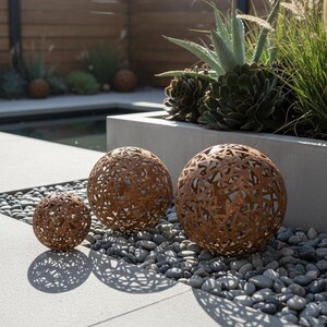 May include: Three decorative metal spheres with intricate cut-out patterns, arranged on a bed of small grey stones. The spheres are various sizes and have a rusty brown colour, casting patterned shadows on the light grey surface.