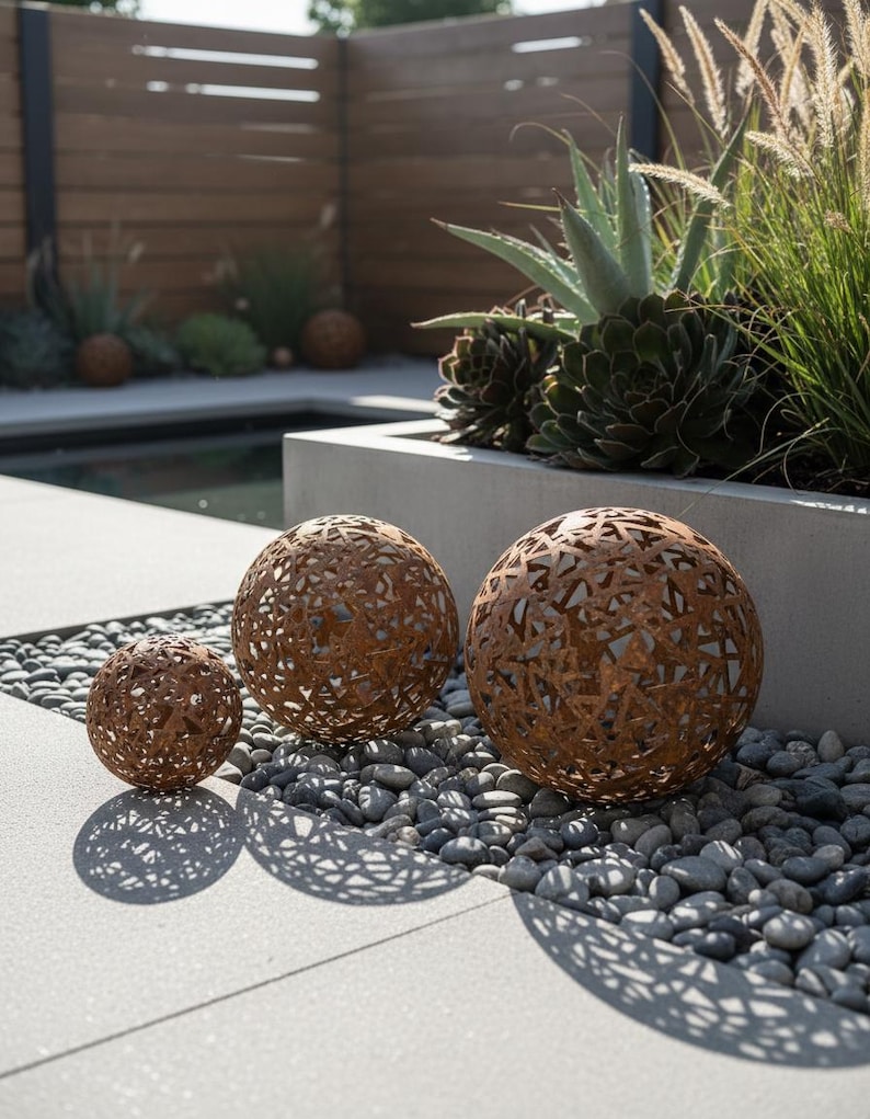 May include: Three decorative metal spheres with intricate cut-out patterns, arranged on a bed of small grey stones. The spheres are various sizes and have a rusty brown colour, casting patterned shadows on the light grey surface.