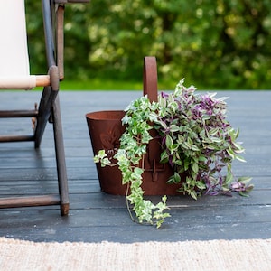 Könnte beinhalten: Ein rostbrauner Metallpflanztopf mit Henkel, gefüllt mit hängenden grünen und lila Pflanzen. Der Pflanztopf hat ein dekoratives Blumendesign und steht auf einer dunklen Holzterrasse. Ein Holzstuhl ist teilweise sichtbar.