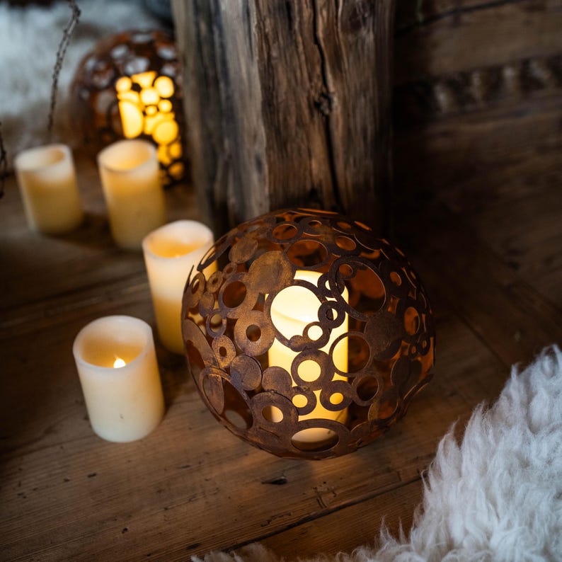 May include: A spherical metal lantern with a warm, glowing light inside. The lantern is made of overlapping, circular metal pieces with a weathered, brown finish. Several battery-operated, ivory-coloured candles are also visible, creating a cosy ambiance.