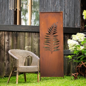 Könnte beinhalten: Ein rechteckiger, brauner Metallschirm mit einem ausgeschnittenen Farnblatt-Design. Der Schirm steht auf einem grünen Rasen neben einem Korbstuhl. Im Hintergrund ein Holzzaun und weiße Blumen.